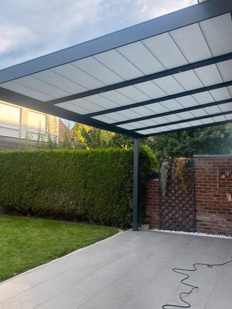 Terrassenüberdachung aus Anthrazit-Aluminium mit Stegplatten, im Abendlicht vor einer hohen Hecke